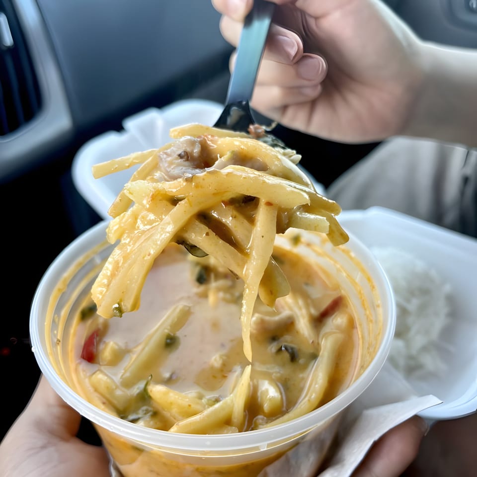 A person holds a clear plastic container of Thai curry with bamboo shoots, taking a spoonful.